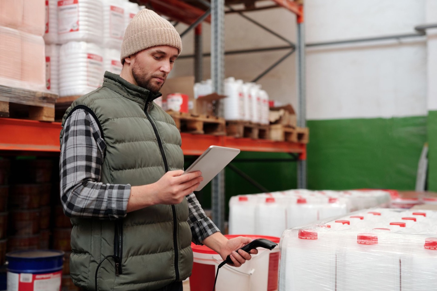 A warehouse manager in a padded vest reading a tablet in a racked aisle, a handheld scanner in his other hand